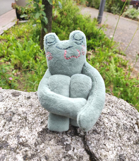 Green frog plushie hugging his legs, content face embroidered on
